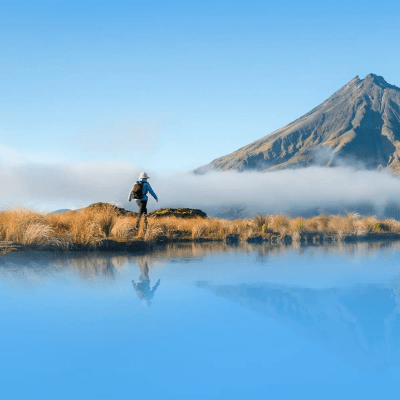 A person with a backpack stands near tall grass by a calm lake, with a mountain and low-lying clouds in the background