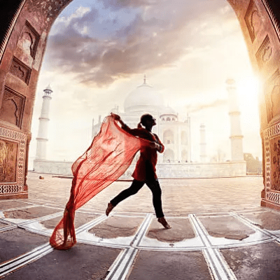 A person holding a red scarf walks through an archway with the Taj Mahal and a dramatic sky in the background