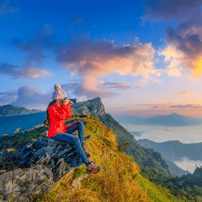 Woman sitting on a mountain peak, gazing at a vibrant sunset with hues of orange and purple in the sky