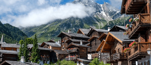 brown wooden houses near green trees and mountain under white clouds during daytime