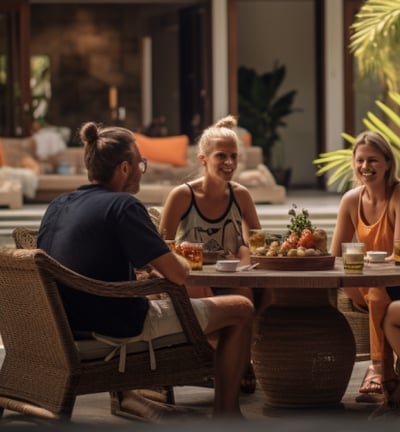 Four people enjoying a meal at a table in a lush tropical setting, surrounded by palm trees and vibrant flowers.