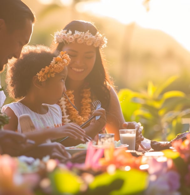 A family enjoying a meal outdoors in Hawaii, with a focus on a child and an adult wearing flower lei
