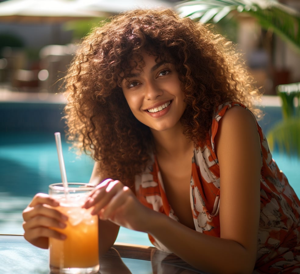 Woman with curly hair sitting poolside, holding orange drink.