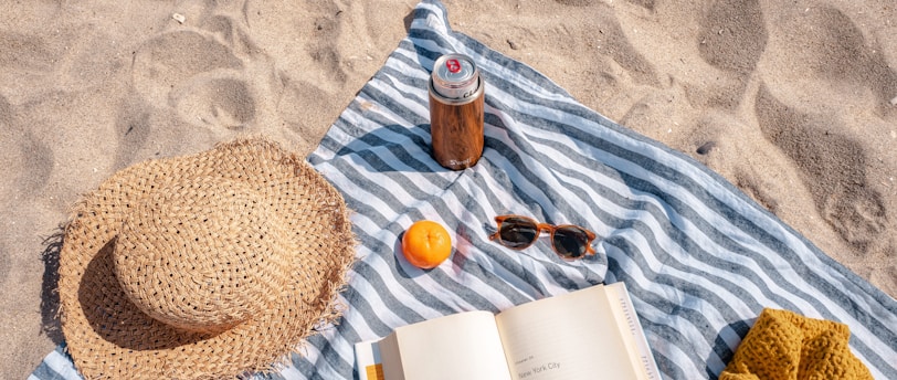 A book, hat, sunglasses, and a straw hat on the beach.